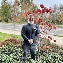 Man with a long beard and a big smile standing in the bushes next to a small tree 