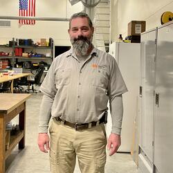 Man with salt and pepper hair, long beard, light brown button up shirt with a long sleeve t-shirt under. 