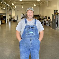 Kevin standing in the shop in blue overalls with his hands in his pockets. 